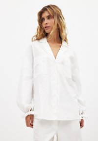 Balta, ilgomis rankovėmis marškiniai, su dviem priekinėmis kišenėmis, laisvas kirpimas, rankogalių juostos ir tekstūruota medvilninė medžiaga.