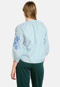 Blusa in cotone a righe con maniche a palloncino, ricamo blu sulla manica sinistra, scollo rotondo e vestibilità comoda, abbinata a pantaloni verdi.
