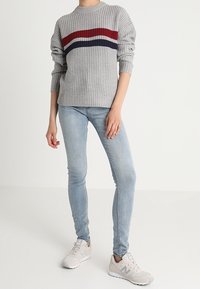 Grau gestrickter Pullover mit roten und marineblauen horizontalen Streifen, kombiniert mit hellblauen Skinny Jeans und cremefarbenen Sneakers.