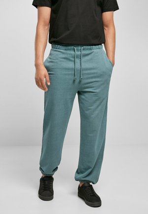 Pantaloni de jogging de culoare teal deschis, confecționați dintr-un material moale, cu un elastic în talie și șnururi, precum și cu mansete elastice, purtați cu adidași negri.