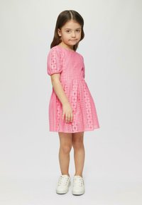Robe en dentelle rose avec des manches courtes bouffantes, dotée de motifs texturés et d'une jupe évasée ; associée à des baskets blanches.