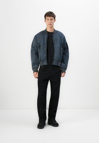 Giovane uomo in piedi con espressione neutra, indossa una giacca bomber blu scuro, una maglietta nera, pantaloni neri e sneakers nere su uno sfondo bianco semplice.