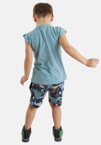 Hellblauses Kurzarmhemd, gemusterte Camouflage-Shorts in Blau, Braun und Beige, schwarze Schuhe mit grünen Akzenten. Kind von hinten gezeigt.