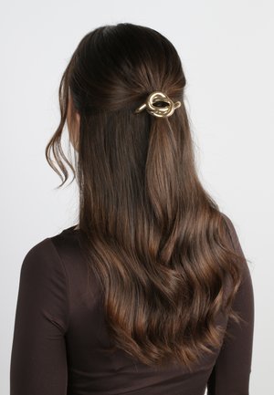 Clip per capelli dorato con design a nodo, che tiene indietro capelli castani mossi. La texture è liscia e lucida, in contrasto con l'abbigliamento scuro.
