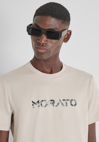 Camiseta beige con el logo "MORATO" en un patrón de mármol. Acompañada de gafas de sol negras y rectangulares. Diseño simple con cuello redondo.
