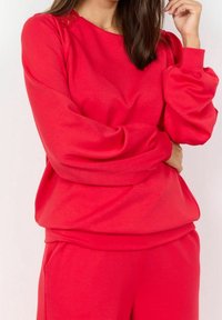 Camiseta de punto roja hecha de una tela suave con cuello redondo y mangas abullonadas, que presenta detalles de encaje en los hombros y un corte relajado.
