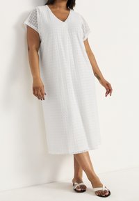 Kvinna iklädd en vit, kortärmad, texturerad midiklänning och vita sandaler, stående med korsade ben mot en enkel vit vägg.