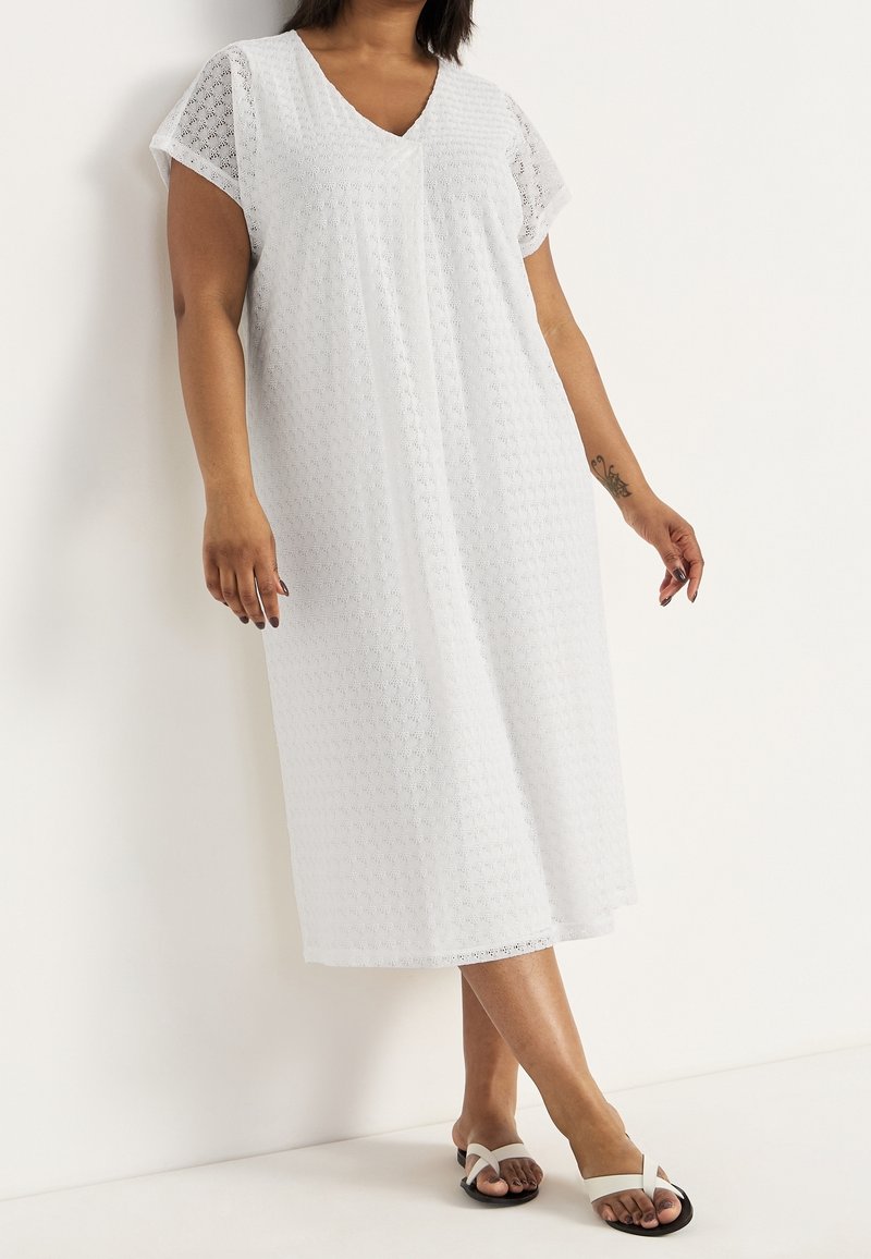 Kvinna iklädd en vit, kortärmad, texturerad midiklänning och vita sandaler, stående med korsade ben mot en enkel vit vägg.