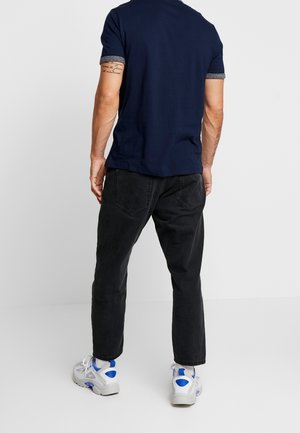 Mężczyzna w granatowej koszulce polo, czarnych dżinsach i białych trampkach z niebieskimi akcentami, stojący tyłem na tle jednolitego, jasnego tła.