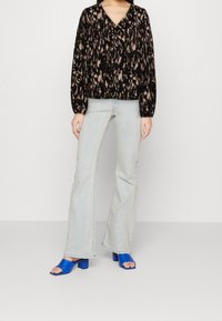 Schwarz-beige gemusterte Bluse mit langen Ärmeln und V-Ausschnitt, kombiniert mit hellblauen, ausgestellten Jeans und blauen Sandalen mit hohem Absatz.