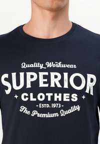 Navyblauwe katoenen t-shirt met witte tekst: "Superior Clothes," opgericht in 1973, met decoratieve sterren en gebogen accenten.