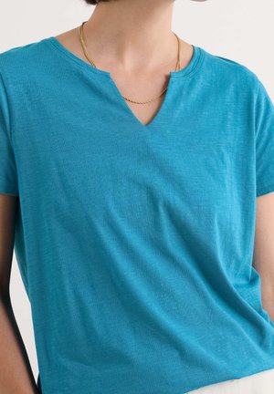 Persoon die een turquoise top met korte mouwen en een gespleten halslijn draagt, met een dunne gouden ketting, tegen een egale achtergrond.