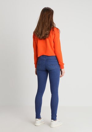 Kvinde iført en orange langærmet top, blå skinny jeans og hvide sneakers, stående med ryggen mod kameraet mod en ensartet lysegrå baggrund.