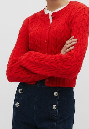 Personne portant un cardigan rouge en maille torsadée boutonné sur un haut blanc, assorti à un pantalon taille haute bleu foncé avec des boutons argentés.
