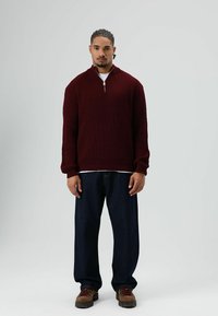 Man met gevlochten haar, gekleed in een donkerrood gebreide trui, losse donkere jeans en bruine wandelschoenen, staand tegen een eenvoudige lichte achtergrond.