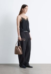 Schwarzes, mit Spitze versehenes Seiden-Camisole, kombiniert mit lockeren schwarzen Hosen und einer braunen Satin-Eimer-Tasche. Das Model trägt schwarze Schuhe und silberne Armbänder.