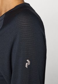 Maglia a maniche lunghe blu navy con texture a costine; presenta un logo ricamato in argento sulla manica. Tessuto morbido e leggero.