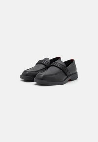 Fekete bőrtalpú, slip-on cipők struktúrált pánttal, amelyen a "HUGO" logó található. Minimalista kialakítás, alacsony sarokkal és piros belső díszítésekkel.