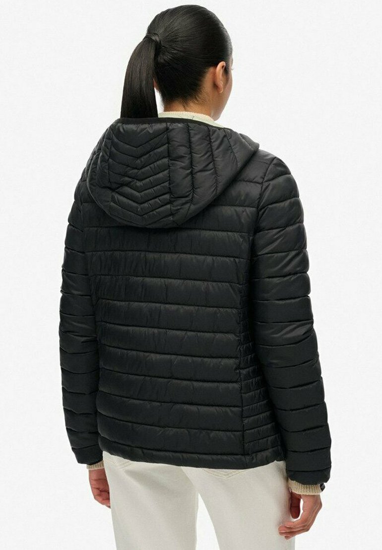 Donna con coda di cavallo nera che indossa una giacca a vento nera trapuntata con cappuccio e pantaloni di colore chiaro, vista da dietro.