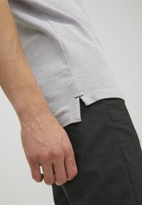 Grå bomullströja med sidöppningar, visad med en hand vilande på mörkt texturerade byxor. Synliga sömnadsdetaljer i accent.