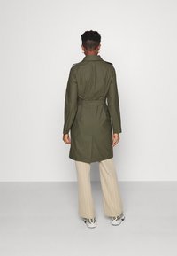 Oliven grøn trenchcoat med bælte, lange ærmer og skulderepauletter, iført over beige stribede bukser og mønstrede sneakers.