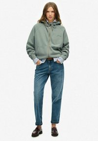 Sweatshirt zippé vert clair avec capuche, doté d'un cordon de serrage et de poches avant, porté par-dessus une chemise rayée bleu et blanc et un jean bleu.