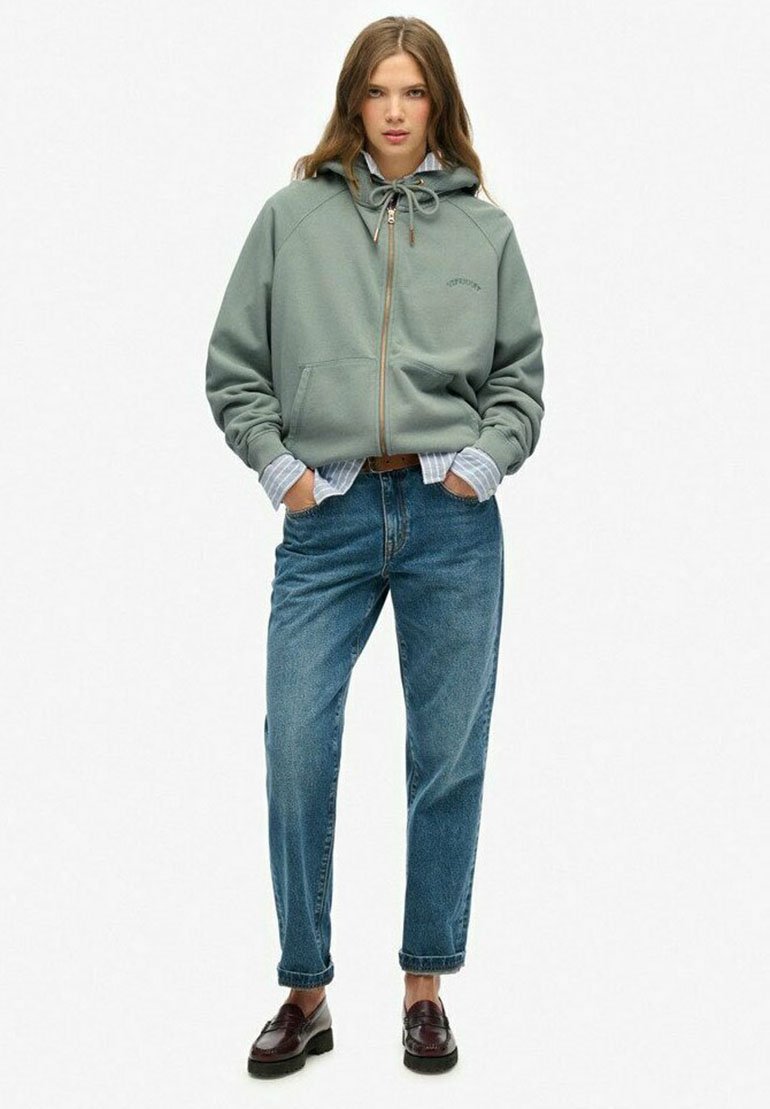 Sweatshirt zippé vert clair avec capuche, doté d'un cordon de serrage et de poches avant, porté par-dessus une chemise rayée bleu et blanc et un jean bleu.