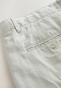 Pantalon beige à la taille avec passants de ceinture, poche fendue à l'arrière et fermeture par bouton, mettant en valeur la texture du tissu et les détails de couture.