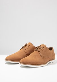 Chaussures en cuir suédé marron à lacets avec une texture lisse, bout arrondi et semelle en caoutchouc blanche. Présentent des coutures simples et un design classique.