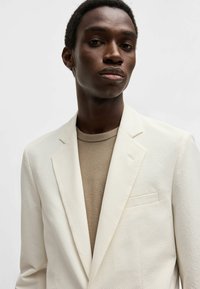 Blazer bianco testurizzato su una maglietta beige a girocollo, con rever a punta, dettagli sottili nelle tasche e una vestibilità strutturata.