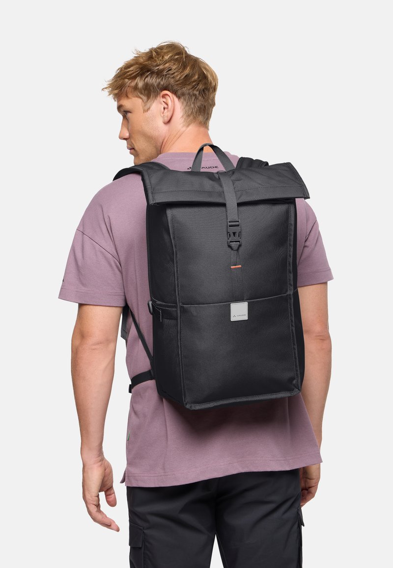 Homme portant un grand sac à dos à fermeture roulante noire avec une boucle avant, vêtu d'un t-shirt mauve à manches courtes et d'un pantalon noir, faisant face à gauche.
