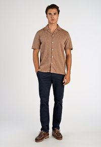 Braunes Kurzarmhemd mit texturierter Stoffstruktur, weißen Knöpfen, marineblauer Hose und braunen Bootsschuhen mit Schnürsenkeln und dicker Gummisohle.