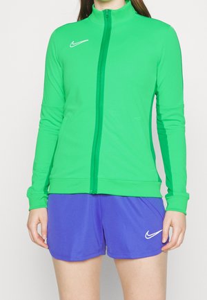 Osoba ubrana w zieloną kurtkę zapinaną na zamek i fioletowe spodenki Nike, stojąca z rozluźnionymi ramionami wzdłuż boków na jednolitym białym tle.