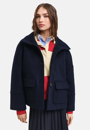 Marineblauwe wollen jas met een hoge kraag, twee voorzakken en een enkele knoopsluiting. Gelaagd over een rood en beige gestreept shirt.