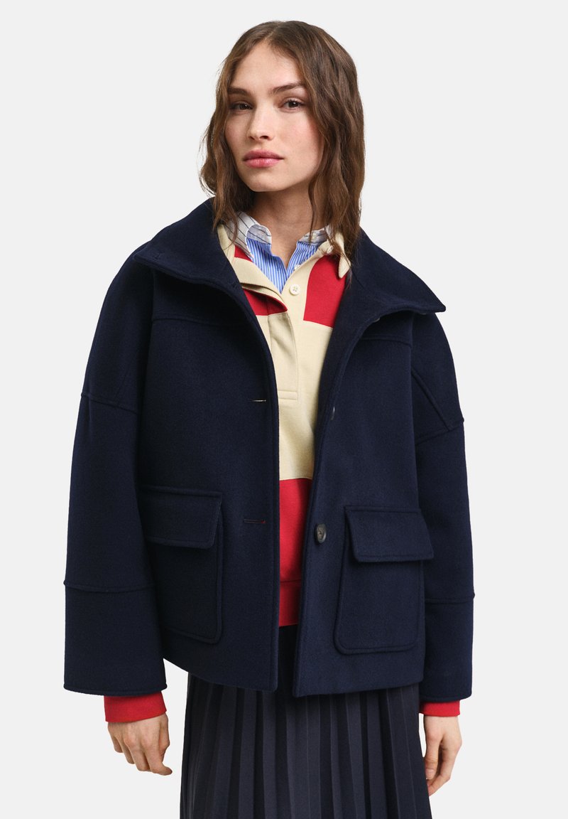 Ναυτικό μάλλινο σακάκι με ψηλό γιακά, δύο μπροστινές τσέπες και κούμπωμα με έναν μόνο κουμπί. Φορεμένο πάνω από ένα κόκκινο και μπεζ ριγέ πουκάμισο.