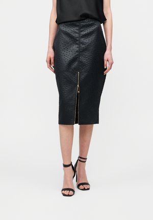 Kvinde iført en sort tekstureret midi-nederdel med en lynlås foran og sorte højhælede sandaler, stående mod en ensfarvet baggrund.