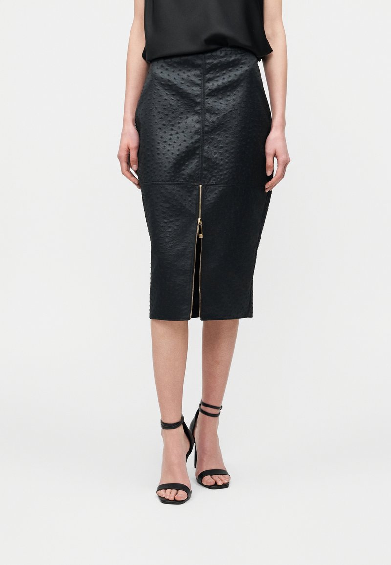 Ženska v črni teksturirani midi krilu z zadrgo spredaj in črnimi sandali z visokimi petami, stoji ob enobarvnem ozadju.