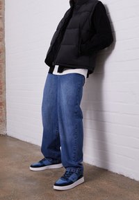 Gilet imbottito nero sopra una camicia bianca, abbinato a jeans blu larghi e sneakers blu con accentuazioni nere, di fronte a un muro bianco.