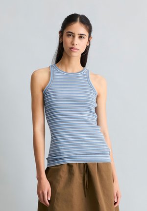 Femme portant un débardeur sans manches bleu et gris rayé et un pantalon marron ample, debout devant un fond uni gris clair.