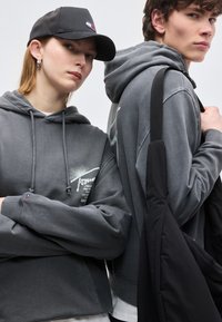 Δύο νέοι που φορούν γκρι φούτερ με κουκούλα· ο ένας φοράει μαύρο καπέλο και ασημένια σκουλαρίκια κρίκους, ο άλλος κουβαλάει μια μαύρη τσάντα ώμου.