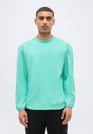 Junger Mann mit kurzen Haaren, der ein helles türkisfarbenes Langarmshirt und schwarze Hosen trägt, steht vor einem einfarbigen hellen Hintergrund.