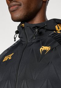 Μαύρο μπουφάν με φερμουάρ και υφασμένο μοτίβο, με κουκούλα, ρυθμιζόμενα κορδόνια και κίτρινο λογότυπο UFC και λεπτομέρειες.