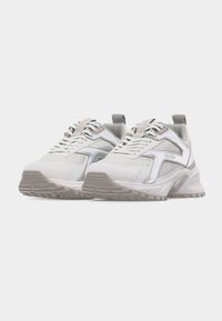 Paire de baskets de sport blanches et argentées avec semelles texturées et design à lacets sur un fond clair uni.