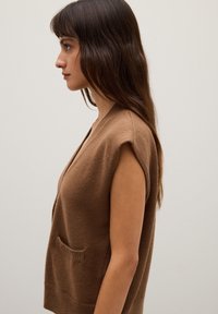 Braune, gestrickte ärmellose Strickjacke mit V-Ausschnitt, ausgestattet mit einer Fronttasche und weichem Material. Minimalistisches Design vor einem neutralen Hintergrund.