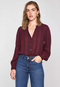 Blouse rayé bordeaux avec un col en V, fermeture à boutons et manches longues bouffantes, associée à un jean bleu taille haute. Texture de tissu lisse.
