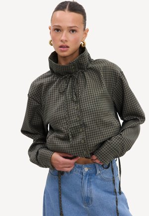 Femme portant une chemise à carreaux verte à col montant boutonnée avec cordons et un jean bleu, posant devant un fond clair uni.
