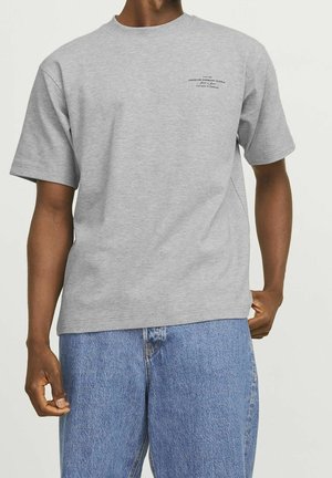 Homme portant un t-shirt gris clair à manches courtes avec un petit texte noir sur la poitrine gauche et un jean en denim bleu, debout les mains détendues.