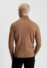 Maglione a collo alto marrone realizzato in tessuto morbido, con un design aderente e polsini a coste, visto da dietro.
