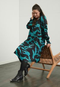 Šaty s čierno-tyrkysovým abstraktným vzorom, dlhými rukávmi s naberacími manžetami a voľnou siluetou s dĺžkou po kolená. Nosené s čiernymi kotníkovými čižmami.