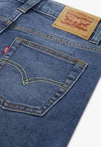 Plave traperice s stražnjim džepom ukrašenim žutim brodskim šavom i kožnom etiketom. Tkanina ima teksturirani tkanje i blago oštećenje.